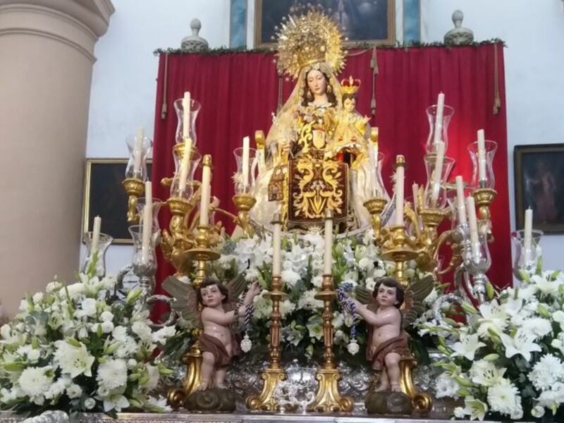Altar con imagen de la Virgen María y dos ángeles, adornado con flores blancas y candelabros dorados.