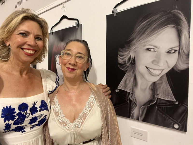 Dos mujeres sonríen junto a una exposición de retratos en blanco y negro.