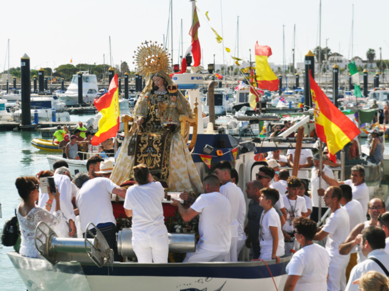 Procesión marítima con la Virgen del Carmen en un barco, rodeada de personas en blanco y banderas españolas en un puerto.