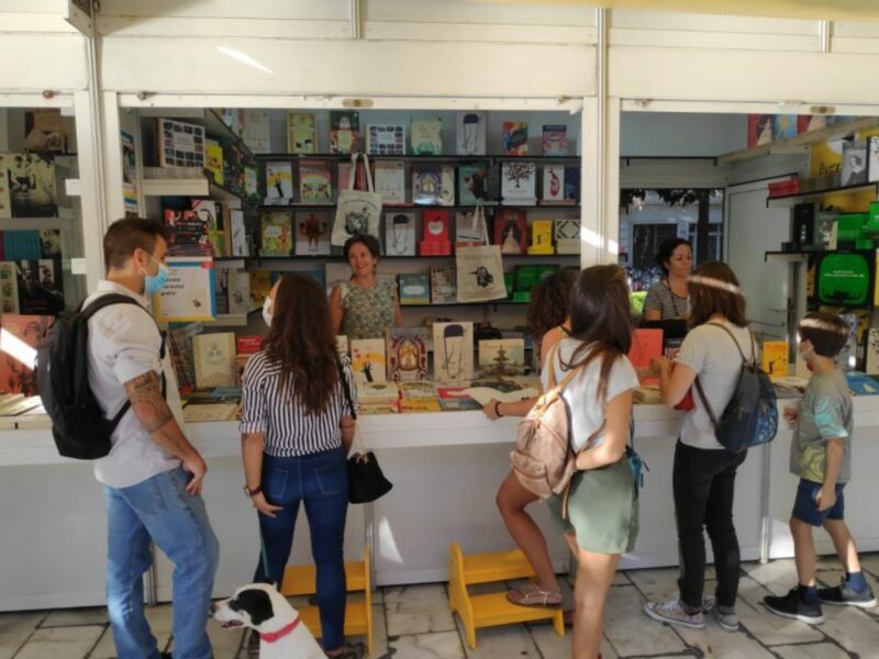 Visita a un puesto de libros en una feria o evento cultural, donde personas de todas las edades seleccionan y adquieren libros.