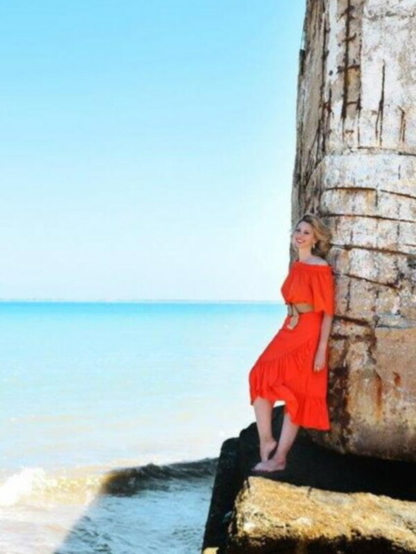 Una mujer con vestido rojo posa sonriendo junto a una roca en la orilla del mar, bajo un cielo azul claro.