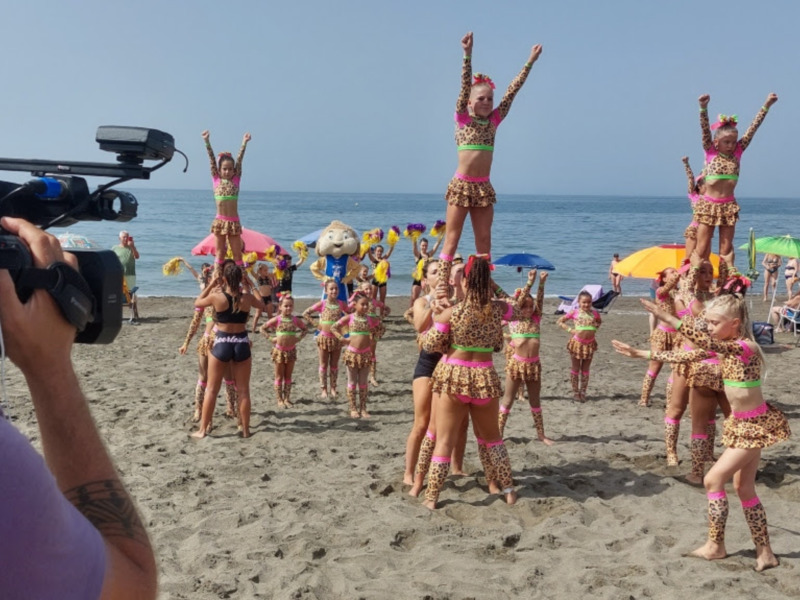 Un grupo de niñas en trajes de leopardo realizando una coreografía en la playa, mientras una persona filmada a la izquierda.