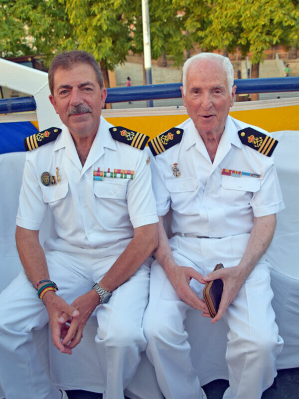Dos oficiales de la mar en uniforme blanco, sentados juntos en un barco, con insignias y medallas en el pecho.