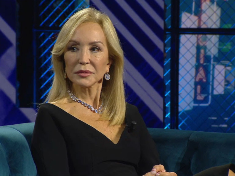 Invitada en un programa de televisión, sentada en un sofá de color azul, con un fondo de estudio con luces y pantallas de televisión.