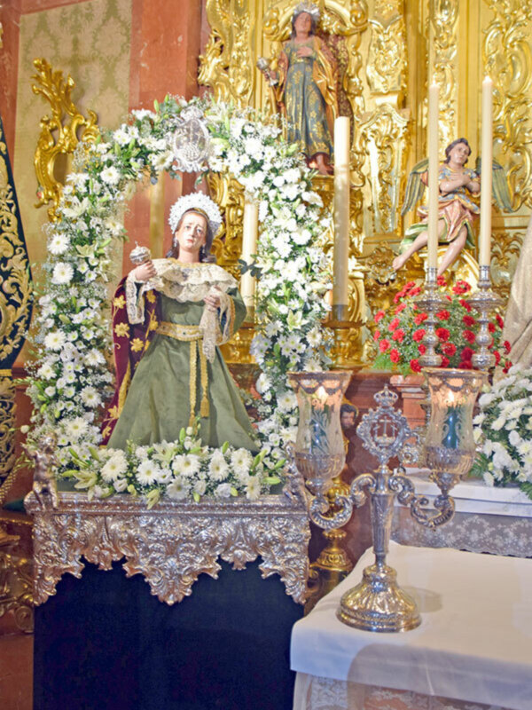 Altar con estatua de la Virgen María en un entorno ornamental y floral, rodeada de velas y adornos dorados.
