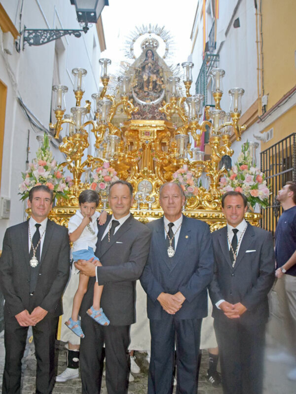 Grupo de personas en trajes formales posando frente a una imagen religiosa dorada y ornamental en una calle estrecha.