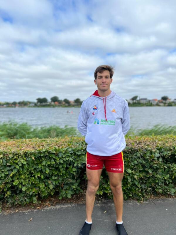 Un atleta posa frente a un lago con un chándal de la selección de Asturias, en un día soleado con nubes dispersas.