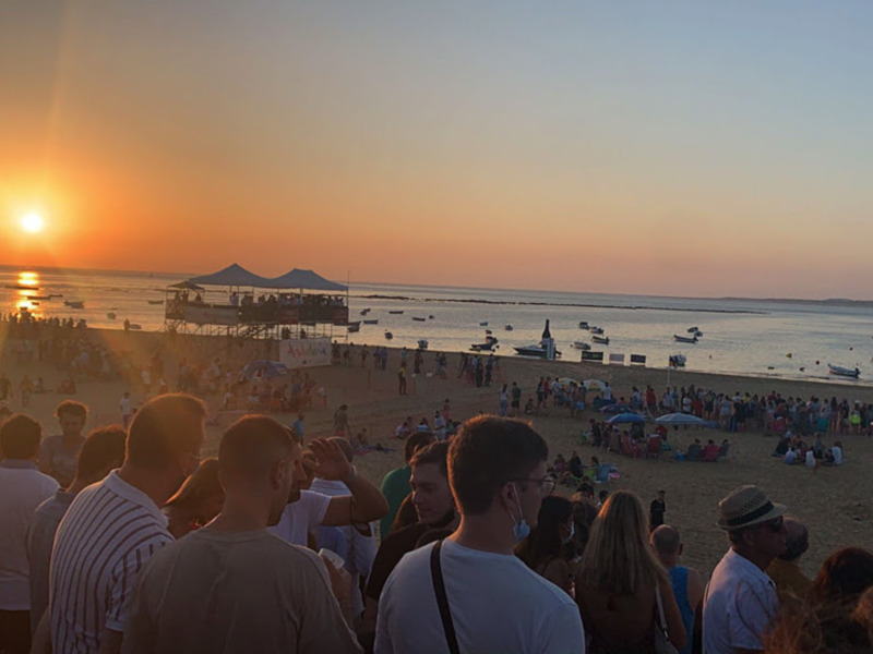 Atardecer en la playa con multitud de personas disfrutando de la vista y el ambiente festivo.