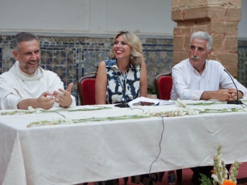 Tres personas sentadas en una mesa con flores y documentos, en un ambiente formal y decorado con azulejos.