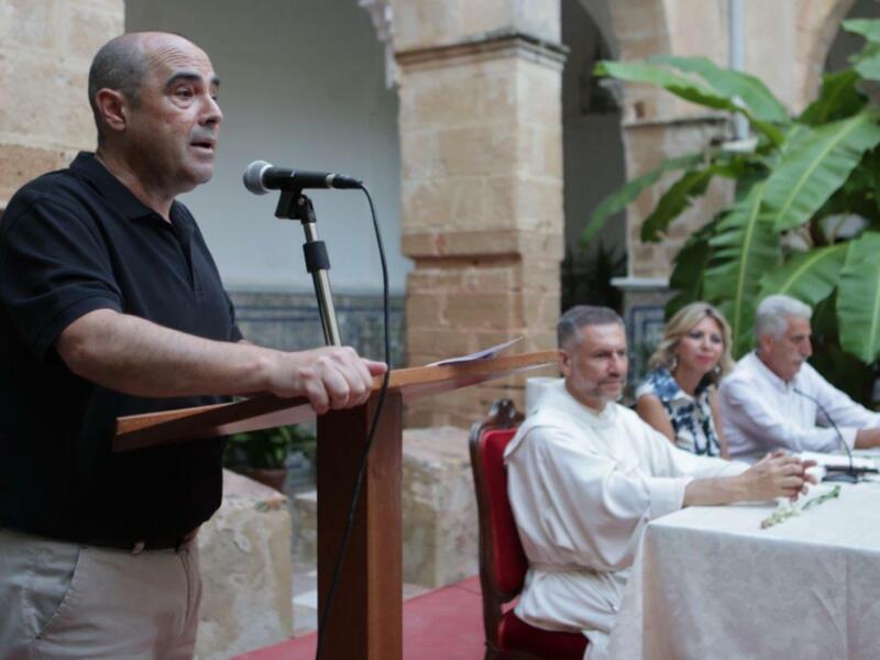 Hombre hablando en un micrófono en un podio, con personas sentadas en la mesa de fondo en un patio histórico.