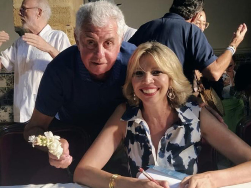 Una pareja en un evento al aire libre, con una mujer sonriendo mientras firma un documento. El hombre la apoya desde detrás.