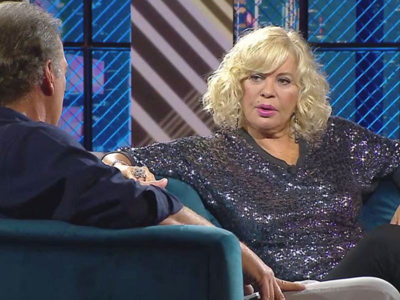 Entrevista en un programa de televisión con una mujer sentada en un sofá, vistiendo un top brillante y charlando con un presentador.