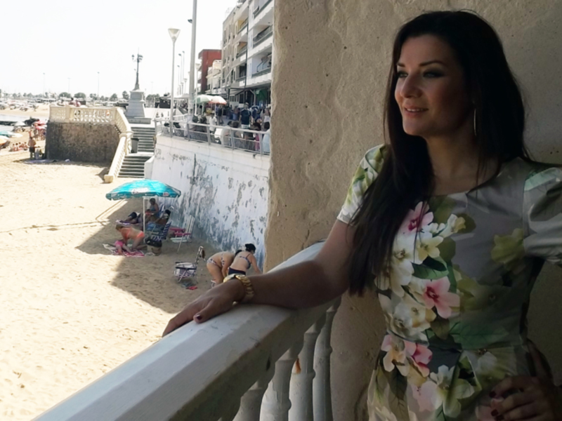 Mujer en vestido floral apoyada en un balcón con vista al mar y playa con sombrillas y bañistas.