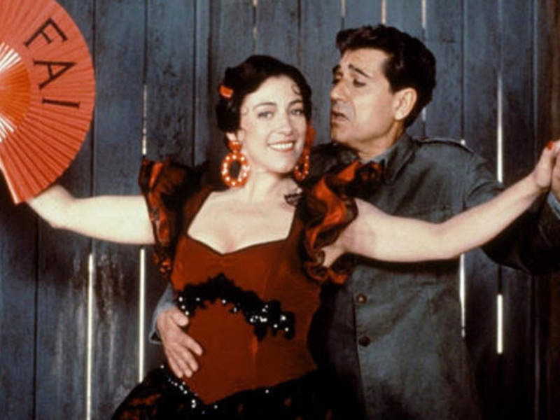 Un par de actores en escena, una mujer con un abanico rojo y un hombre en uniforme, posando con expresiones dramáticas.