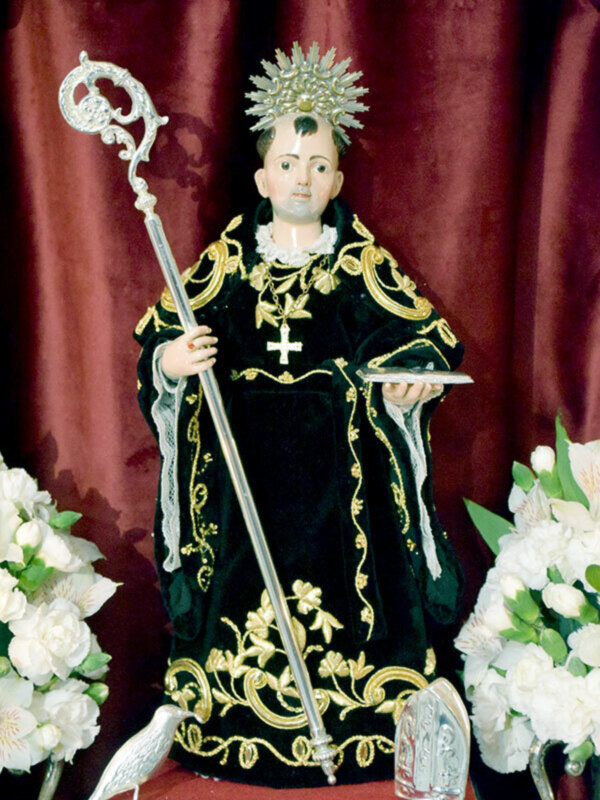 Figura religiosa con traje negro y dorado, manto, bastón y corona, flanqueada por flores blancas.