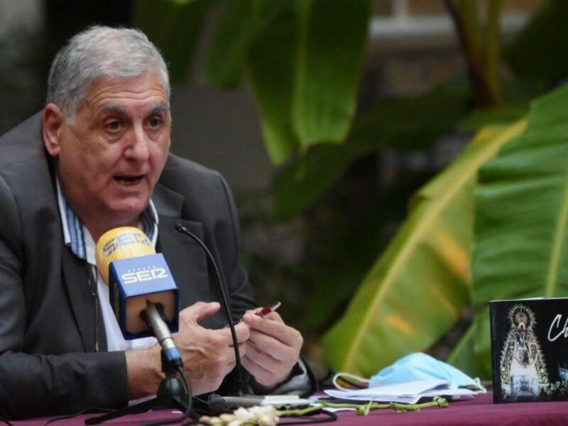 Persona hablando en rueda de prensa con micrófono y libro titulado 'Regla de Chipiona' en la mesa.