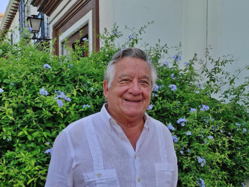 Retrato de hombre sonriente en un jardín con flores azules y edificio blanco en el fondo.