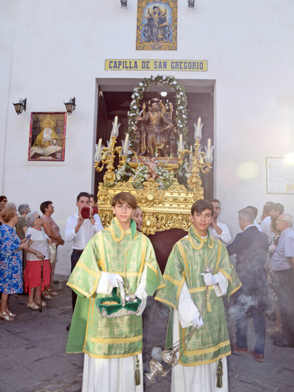 Procesión religiosa frente a la Capilla de San Gregorio, con jóvenes portando candelabros y un altar ornado con flores y velas.
