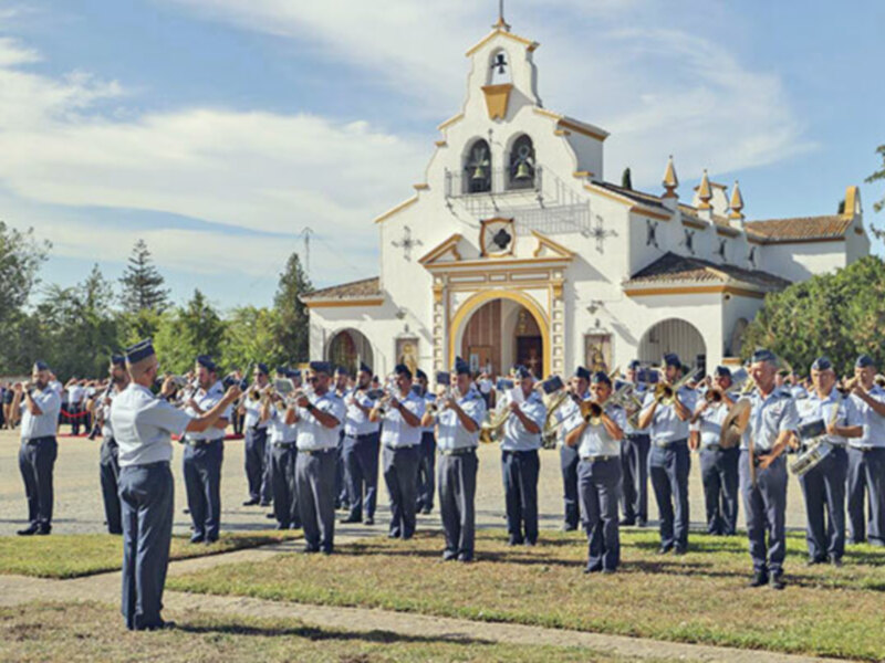 Banda de música en uniforme tocando frente a una iglesia blanca con detalles dorados.