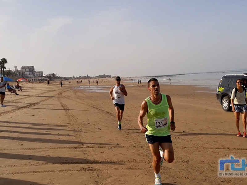 Corredores participando en una carrera de maratón en la playa, con el mar y la ciudad en el fondo.