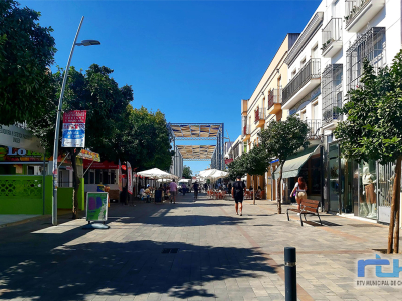 Calle comercial con tiendas y restaurantes, iluminación moderna y árboles en el centro. Imagen de Chipiona, Cádiz.