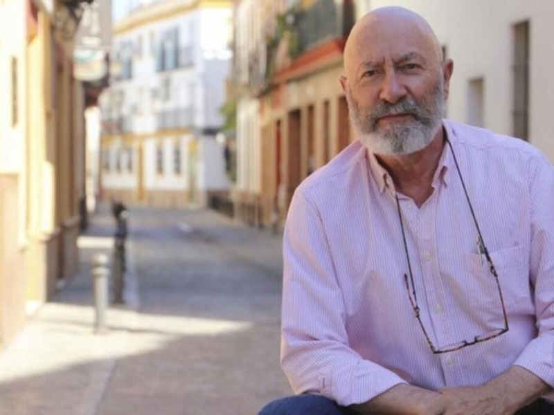 Un hombre con barba y camisa rosa posa en una calle de casas coloridas.