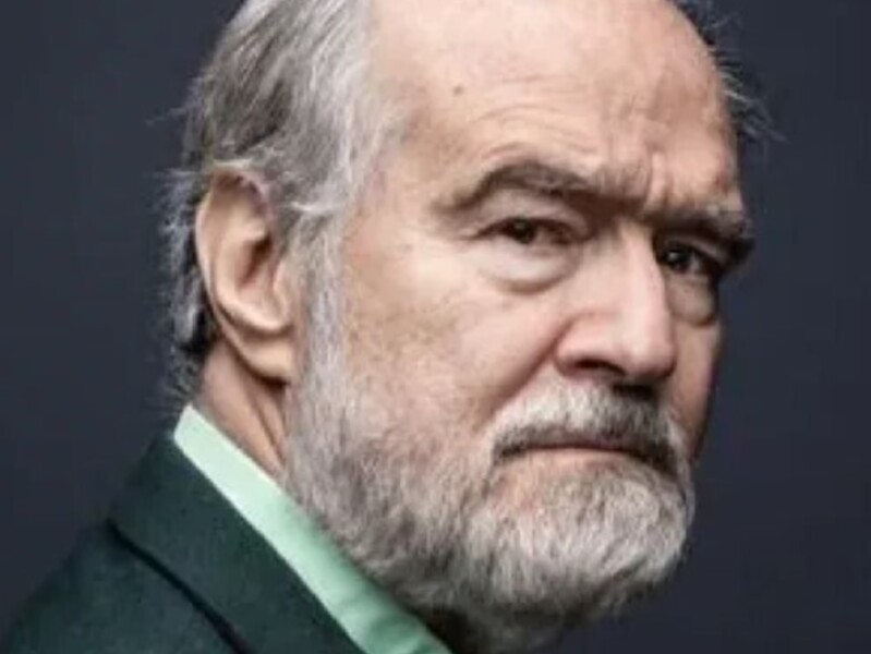 Retrato de hombre maduro con barba y cabello gris, vistiendo un traje oscuro con camisa verde, contra un fondo oscuro.