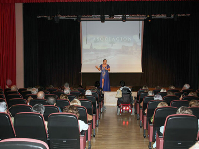 Una mujer en silla de ruedas da una presentación en un auditorio con asistentes sentados en sillas rojas y negras, frente a un proyector con la palabra 'ASOCIACIÓN' en la pantalla.
