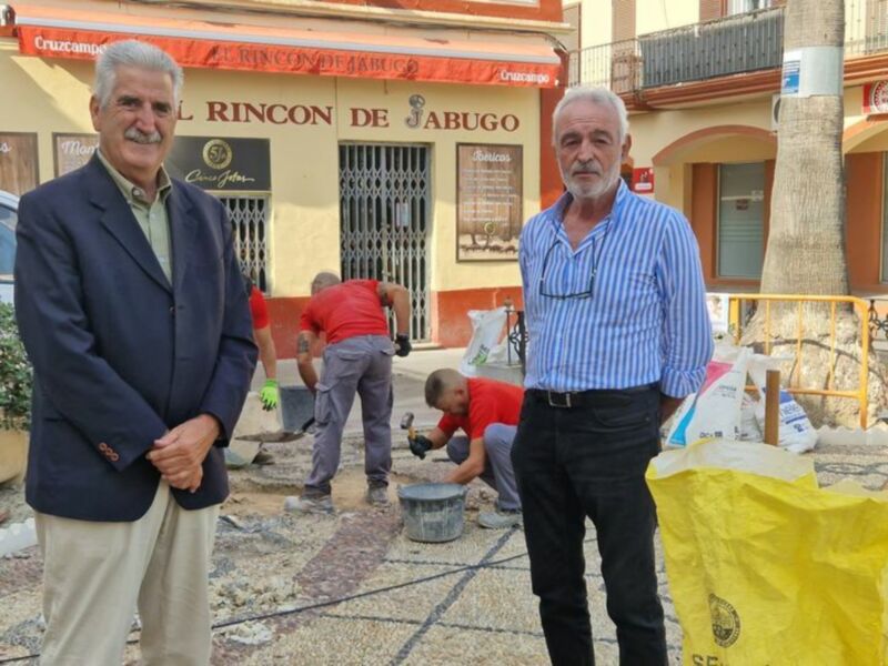 Dos hombres posan frente a trabajadores que reparan una calle en un pueblo con fachada de tienda llamada 'El Rincon de Jabugo'.