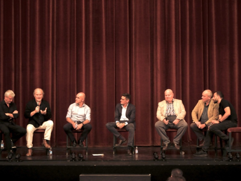 Panel de discusión en escenario con seis participantes sentados en sillas, fondo de cortina roja.
