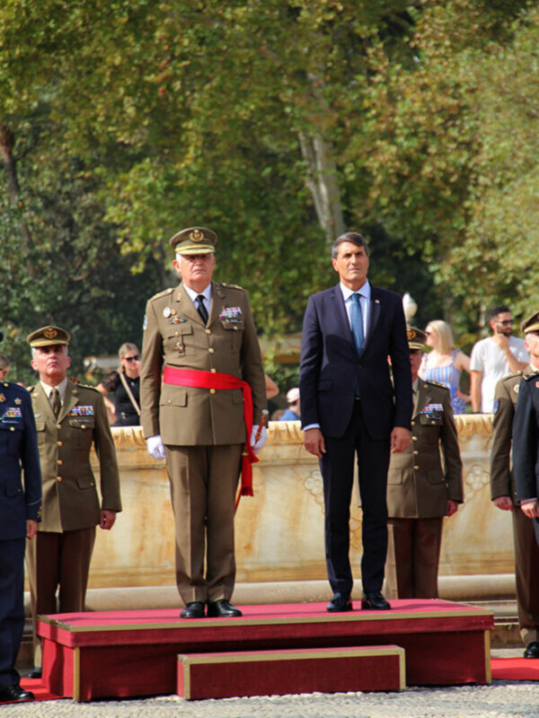 Oficiales militares y civiles en una ceremonia formal al aire libre, con árboles y público en el fondo.