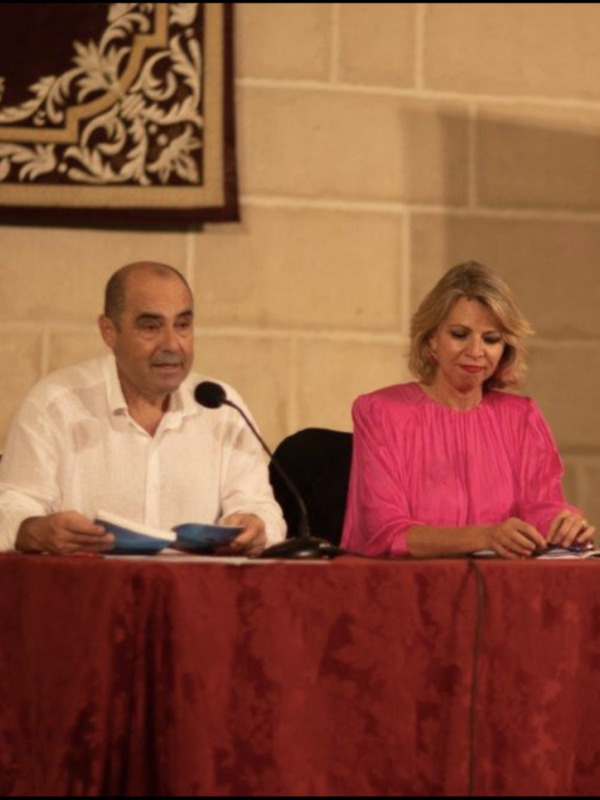 Persona hablando en un evento formal con micrófono y otra persona sentada junto a ella.