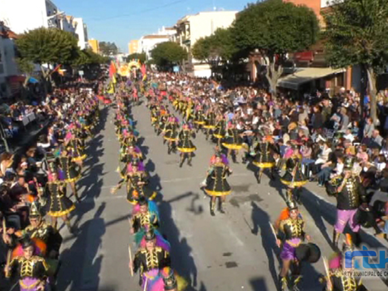 Desfile de carnaval en la calle, con participantes vestidos de colores y disfraces. Público observando desde los lados.