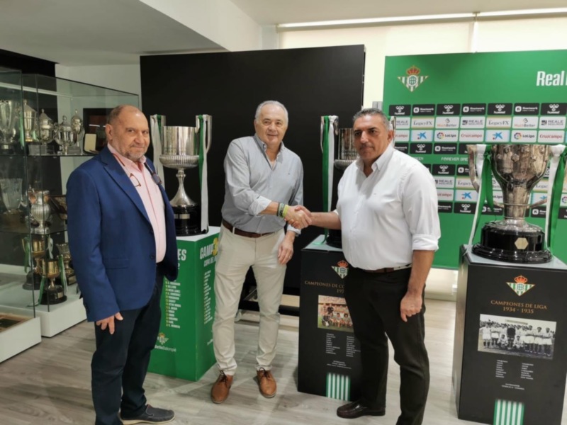 Dos hombres saludándose en una sala con trofeos y banderas de equipos deportivos en el fondo.