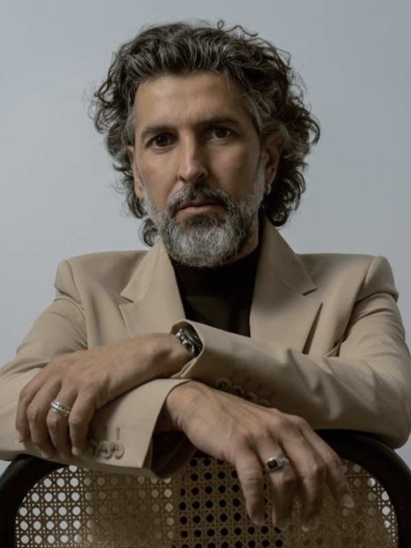 Retrato de hombre con cabello rizado y barba, vestido con traje beige y sentado en una silla de madera con patrón geométrico.
