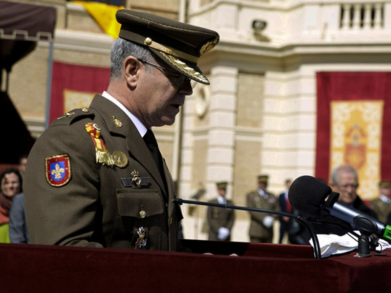 Un oficial militar habla en un acto oficial en un escenario con banderas y edificios históricos al fondo.