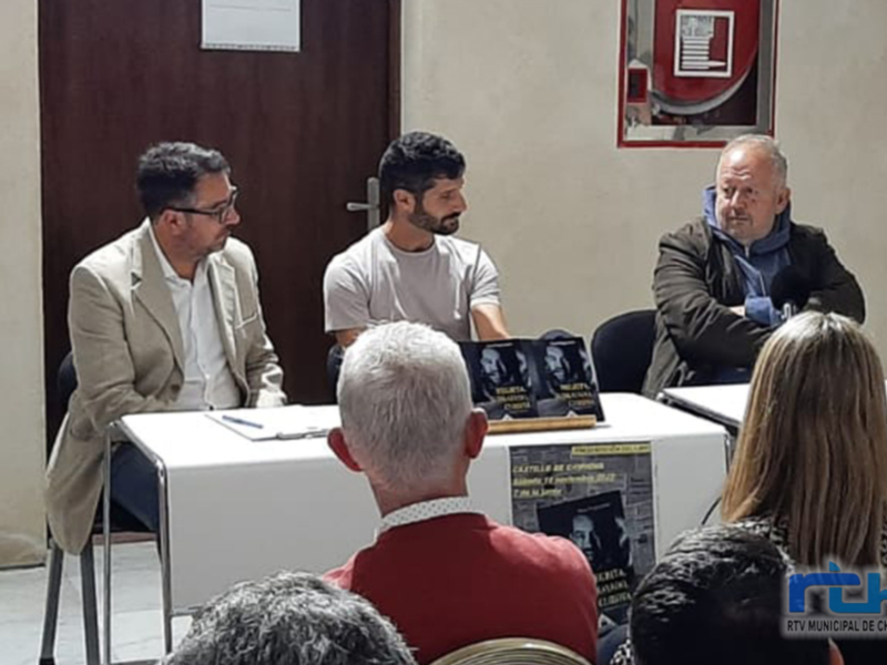 Entrevista en un evento público, tres personas sentadas en una mesa con audiencia en primer plano.