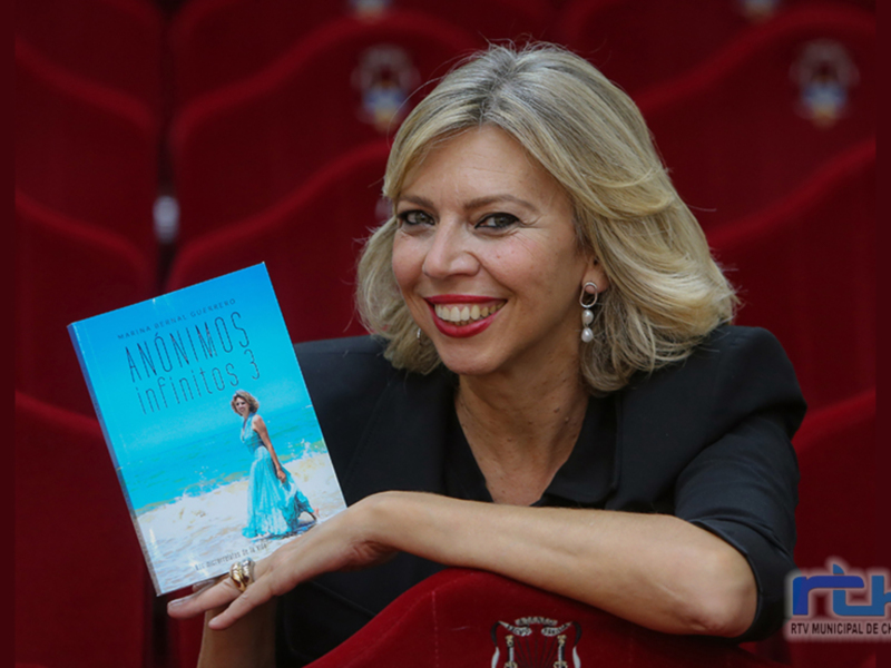 Una mujer sonriente sostiene un libro titulado 'Anónimos Infinitos 3' frente a un fondo rojo con un logo.