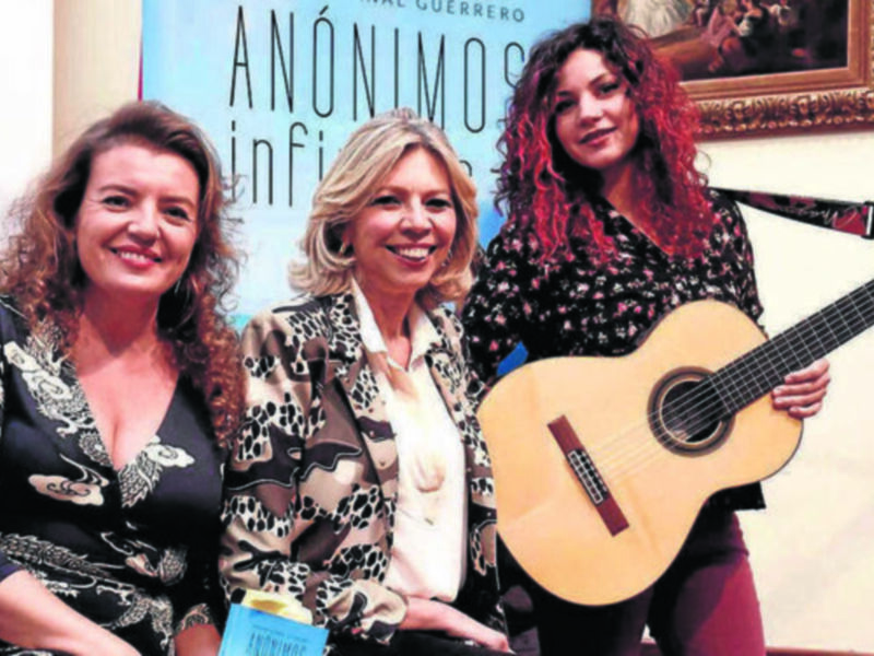 Tres mujeres sonríen juntas, una toca una guitarra clásica, con un cartel de 'Anónimos' en el fondo.