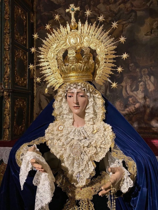 Imaginaria de la Virgen María en una iglesia, vestida con una capa azul y un manto dorado, con una corona de estrellas y un halo dorado. La imagen se encuentra en un entorno religioso con un fondo que muestra escenas de la vida de Cristo.