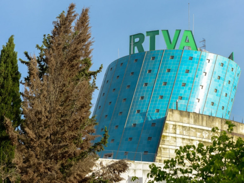 Torre azul con letras grandes 'RTVA' en el cielo azul claro, rodeada de árboles verdes y un cielo azul claro.