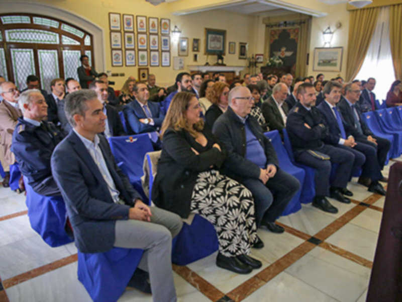 Una audiencia formal en un salón con sillas azules, personas sentadas y un fondo con cuadros y una gran imagen en el centro.