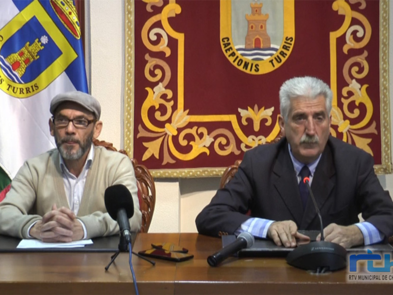 Conferencia de prensa en Chipiona, con representantes municipales sentados en un escritorio con microfonos y banderas oficiales en el fondo.