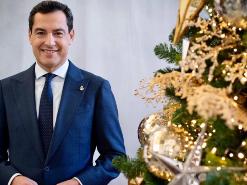 Un hombre en traje azul posa junto a un árbol de Navidad decorado con luces y adornos dorados.