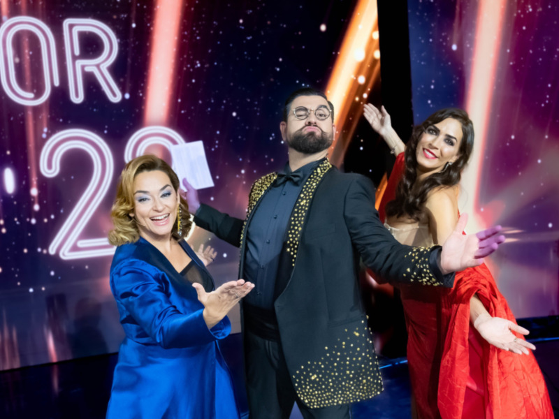Tres personas en trajes elegantes posando en un escenario con luces y el número '29' en el fondo.