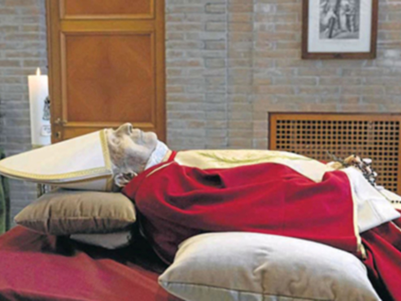 Un altar con una figura vestida con un manto rojo y un tocado dorado, rodeada por un cojín beige y una vela en la esquina. La pared detrás es de ladrillo y el suelo es de baldosas.