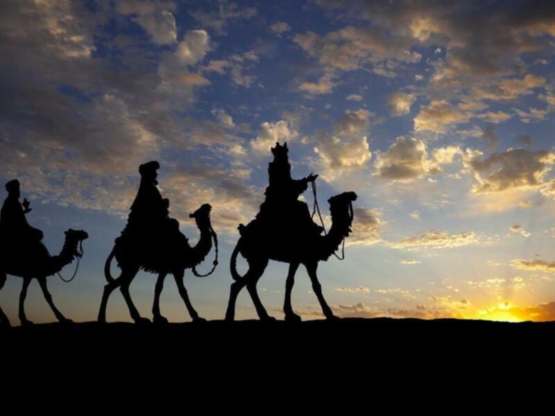 Silueta de tres hombres montando camellos en el desierto, con un cielo dramático y nubes en el fondo.
