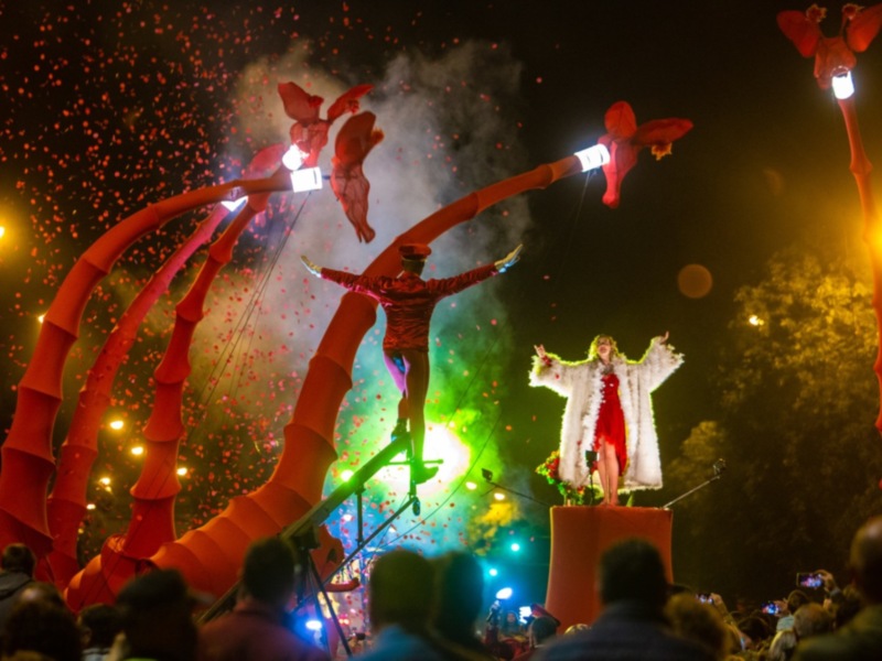 Desfile nocturno con artistas en trampolines y confeti rojo, iluminado por luces coloridas.