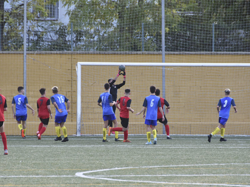Una imagen de un partido de fútbol en curso, con jugadores en dos equipos distintos: uno con camisetas rojas y otro con azules. El portero está en acción, mientras los demás observan el juego desde la portería. La cancha es verde y bien mantenida, con líneas blancas marcando el campo de juego. En el fondo se puede ver una barda y árboles, lo que sugiere un entorno urbano.