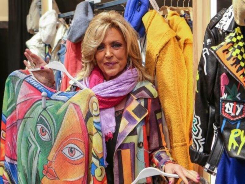 Una mujer elegante con abrigo colorido y bufanda posa en un showroom de ropa, rodeada de prendas vibrantes y modernas.